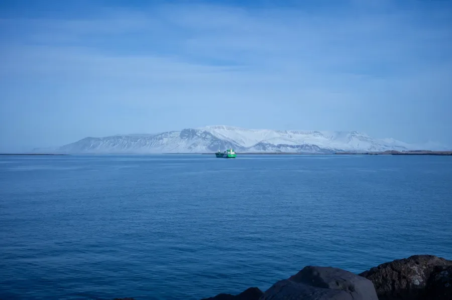A single boat crosses still water at twilight, a snow-covered range low along the far shore. Photographed from the Reykjavík shoreline beside the Sun Voyager, facing the way it points.
